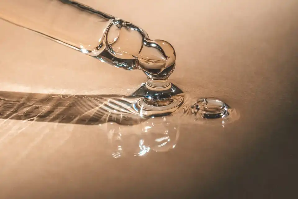 Eine Nahaufnahme einer Glaspipette, die eine klare Flüssigkeit auf eine glatte, reflektierende Oberfläche abgibt. Die Flüssigkeit bildet kleine Pfützen und die Pipette wirft einen detaillierten Schatten. Das Bild hat einen warmen, goldbraunen Ton.