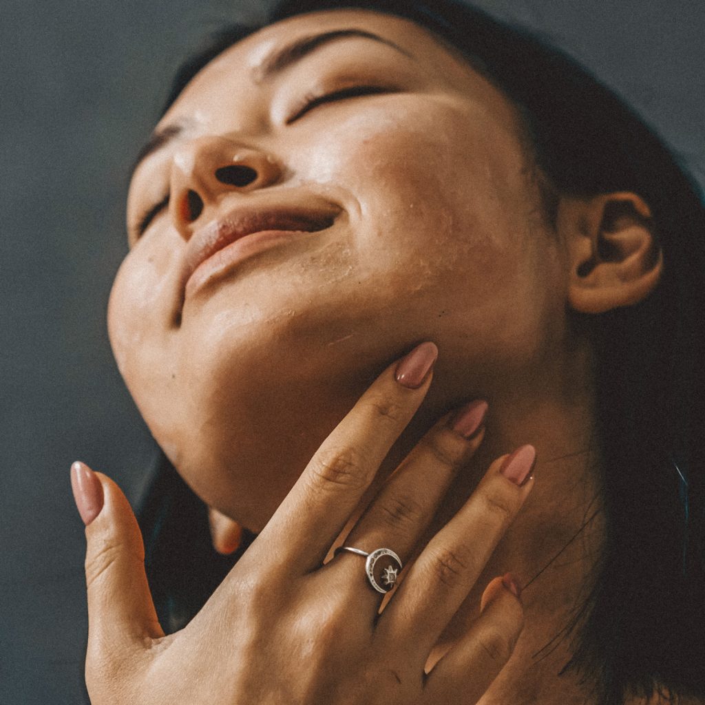 Eine Frau mit langem, dunklem Haar hat den Kopf in den Nacken gelegt, die Augen geschlossen und lächelt sanft. Sie berührt mit der linken Hand ihren Hals und trägt einen silbernen Ring mit einem kleinen Blumenmuster. Der Hintergrund ist dunkel und unterstreicht ihren heiteren Gesichtsausdruck.