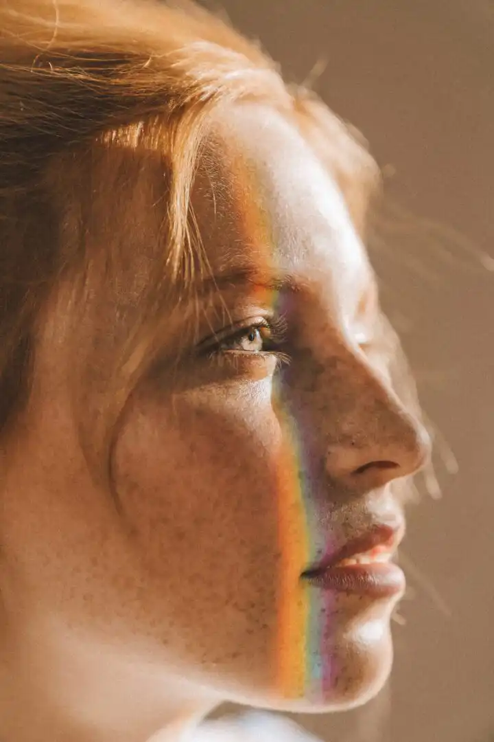 Eine Person mit heller Haut und roten Haaren blickt mit sanftem Gesichtsausdruck zur Seite. Ein Regenbogen aus Licht fällt über ihr Gesicht und hebt ihre Sommersprossen hervor. Der Hintergrund ist verschwommen und hat einen neutralen Ton.
