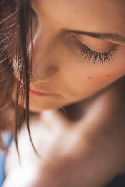 Nahaufnahme einer Person mit langen Wimpern und braunem Haar, das ihr ins Gesicht fällt. Die Person blickt nach unten, wodurch ihre Wimpern und ihre glatte Haut betont werden. Die Beleuchtung erzeugt eine sanfte, intime Atmosphäre.