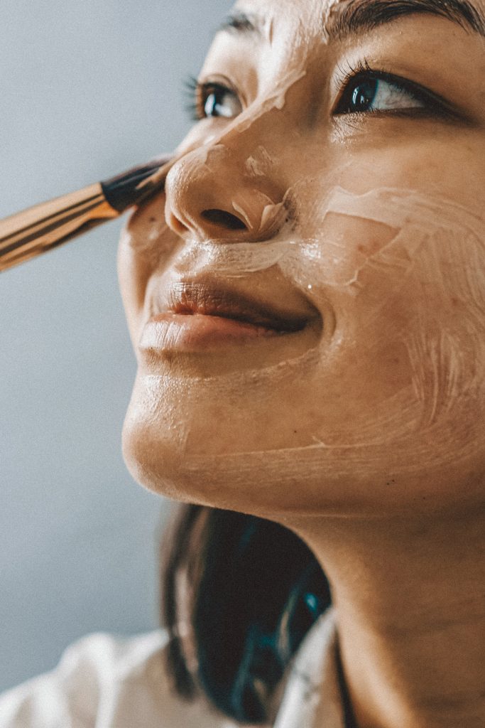 Eine Person lächelt, während sie mit einem Pinsel eine Gesichtsmaske aufträgt. Die Maske wird auf der Wange und um die Augenpartie verteilt. Der Hintergrund ist sanft verschwommen, wodurch der Ausdruck und die glatte Textur der Maske hervorgehoben werden.